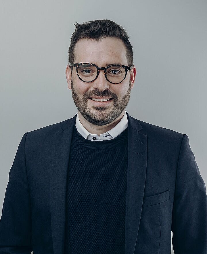 Felix Greiter lächelnd mit Brille und in dunkelblauer Kleidung vor hellem Hintergrund.