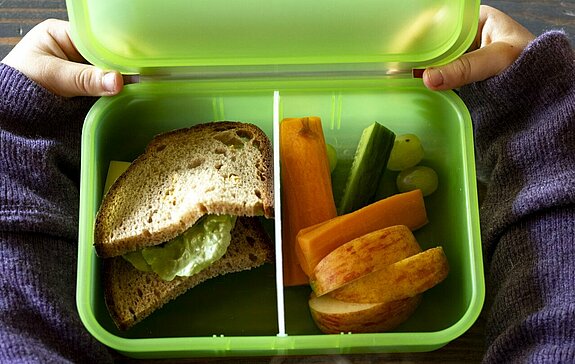 Kind sitzt Tisch vor der ZAK Brotzeitdose. Die Brotzeitdose ist gefüllt mit Gemüse, Obst und einem belegten Brot.