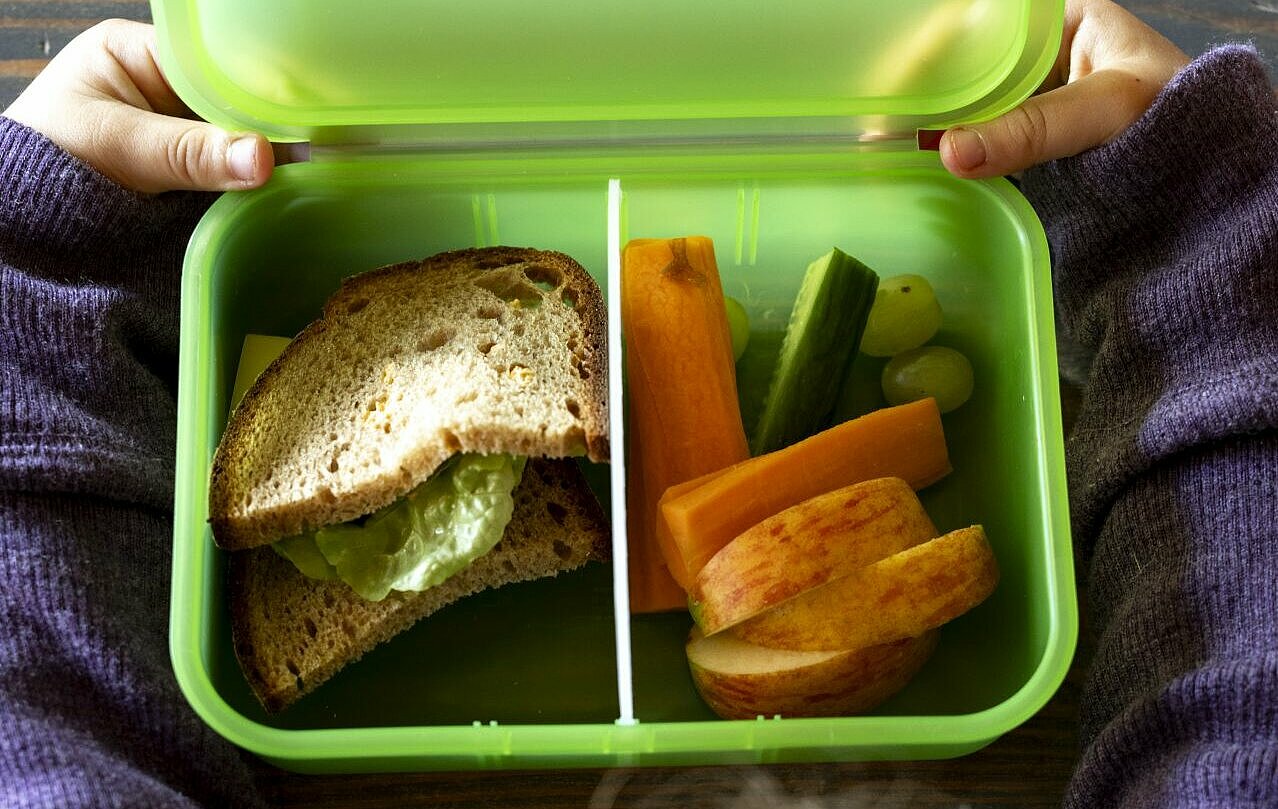 Kind sitzt Tisch vor der ZAK Brotzeitdose. Die Brotzeitdose ist gefüllt mit Gemüse, Obst und einem belegten Brot.