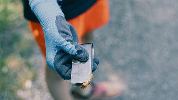 Hand mit blauer Arbeitshandschuh hält eine Metallzange mit einem kleinen Müllstück