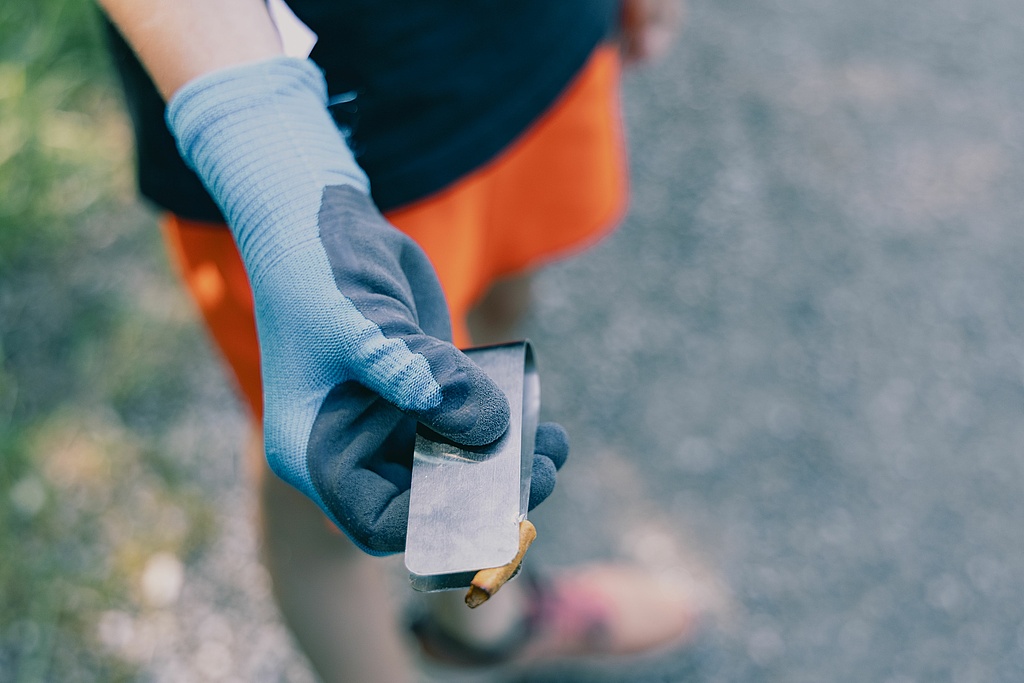 Hand mit blauer Arbeitshandschuh hält eine Metallzange mit einem kleinen Müllstück
